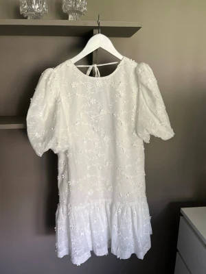 Klänning - Supersöt vit kortklänning med broderade blommor, puffärmar och volangkant. Klänningen har öppen rygg med knytband och är perfekt för sommarens alla tillfällen. Materialet känns lätt och luftigt, och den har en romantisk vibe. Prislappen kvar.