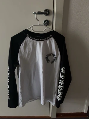 Chrome Hearts svartvit långärmad tröja - Säljer en svartvit långärmad tröja från Chrome Hearts med kontrastärmar och tryck på både bröstet, ryggen och ärmarna. Tröjan har en avslappnad passform och är tillverkad i mjuk bomull. Perfekt för dig som gillar streetwear och tydliga loggor.