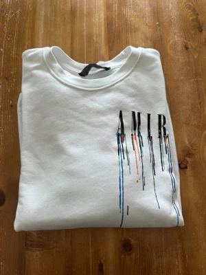 Vit sweatshirt från Amiri - Säljer en vit sweatshirt från Amiri med tryckt logga och färgstänk i blått, rött och svart på bröstet och nedtill. Tröjan har rund halsringning och ribbade muddar vid ärmslut och nederkant. Perfekt för dig som gillar streetwear och unik design.