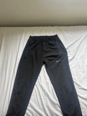 Svarta Nike mjukisbyxor med dragkedja - Säljer ett par svarta mjukisbyxor från Nike med elastisk midja och dragkedjor på fickorna. Byxorna har en smal passform och den klassiska Nike-loggan broderad på benet. Perfekta för chill eller träning, riktigt sköna och stilrena.