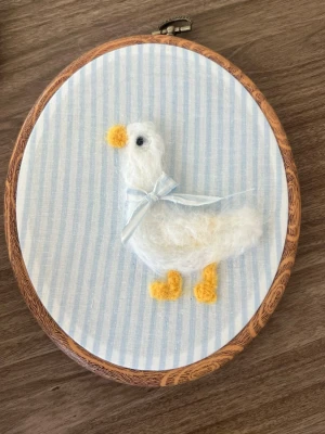 Broderad tavla med filtad gås - Söt handgjord tavla med en vit filtad gås på blå- och vitrandig tygbakgrund. Gåsen har gula ben och näbb samt ett ljusblått sidenband runt halsen. Monterad i en rund träram med metallspänne, perfekt som dekoration.