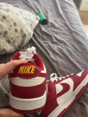 Nike Dunk Low röd och vit sneaker - Nike Dunk Low sneakers i rött och vitt med klassisk Swoosh på sidan och gul NIKE-text på hälen. Ovandelen är i läder med vita och röda paneler, vita skosnören och röd yttersula. Perfekt för dig som vill ha en ikonisk och stilren sneaker.