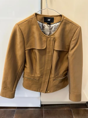 Brun jacka från H&M med dragkedja - Snygg brun jacka från H&M med rund halsringning och guldfärgade dragkedjor. Jackan har långärmade ärmar och ett coolt mönstrat innerfoder. Perfekt för dig som vill ha en stilren och trendig look.