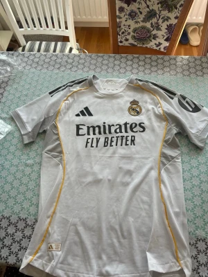 Real Madrid matchtröja Adidas vit - Säljer en officiell Real Madrid matchtröja från Adidas i vitt med svarta detaljer och gula inslag. Tröjan har trycket 'Arda Güler 15' på ryggen och klubbmärke på bröstet. Tillverkad i lätt och ventilerande material, perfekt för fotboll eller träning.