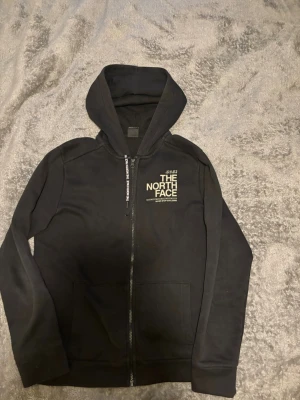 Svart hoodie från The North Face - Klassisk svart hoodie från The North Face med dragkedja framtill och vit logga på bröstet samt ryggen. Hoodien har stora fickor framtill och justerbar huva. Perfekt för en avslappnad och stilren look.