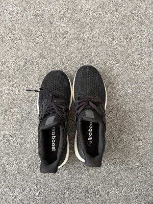 Svarta Adidas Ultraboost sneakers - Säljer ett par svarta Adidas Ultraboost sneakers med stickad ovandel och klassiska tre ränder på sidan. Skorna har vita Boost-sulor och svarta detaljer i plast vid snörningen. Perfekta för dig som vill ha både stil och komfort.
