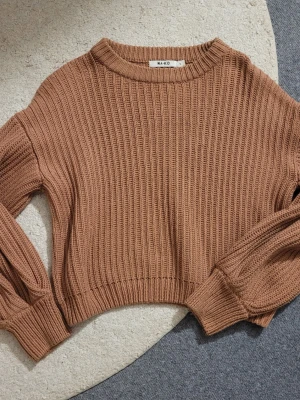 Brun ribbstickad tröja från NA-KD - Säljer en snygg brun ribbstickad tröja från NA-KD med croppad passform och rund halsringning. Tröjan har långa ärmar med breda muddar och ett chunky stickat mönster som ger en trendig look.