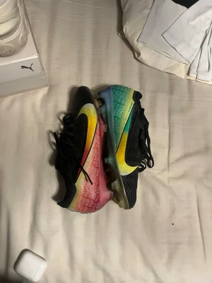 Nike Mercurial vini fotbollsskor - Säljer ett par Nike Mercurial fotbollsskor med svart ovandel och färgglad sula i rött, gult, grönt och blått. Skorna har en stor gul Nike-swoosh på sidan och snörning framtill. Ovandelen är i syntetmaterial med struktur för bättre bollkontroll. Perfekta för dig som vill sticka ut på planen.