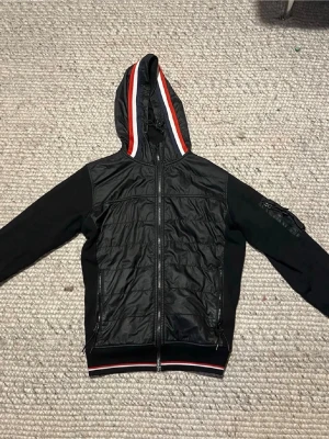 Svart Moncler cardiganjacka med huva - Snygg svart cardiganjacka från Moncler med huva och dragkedja. Jackan har röda och vita ränder på huvan och nederkanten, samt fickor med dragkedja. Materialmix med glansig front och ärmar i mjuk bomull. Moncler-logga på ärmen.