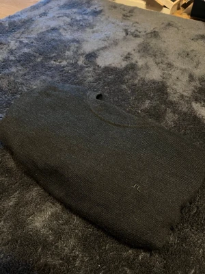 Mörkgrå stickad tröja från J.Lindeberg i merinoull  - Mörkgrå stickad tröja från J.Lindeberg med rund halsringning och diskret logga broderad på bröstet. Tröjan har långa ärmar och ribbade muddar vid ärmslut och nederkant. Perfekt för lager på lager och enkel att matcha.