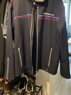 BMW Motorsport svart jacka - Snygg svart jacka från BMW Motorsport med dragkedjor och detaljer i blått och rött. Jackan har ståkrage med knapp, två fickor med dragkedja och sportig design. Perfekt för dig som gillar racing och streetwear. Möts upp i Västerås snabb deal 1500kr