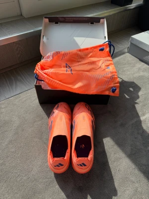 Adidas orange fotbollsskor F50 - Säljer ett par Adidas F50 fotbollsskor i en riktigt snygg orange färg med blå detaljer. Skorna har en strömlinjeformad design och är tillverkade i lätt syntetmaterial för maximal fart på planen. Levereras med matchande orange skopåse.