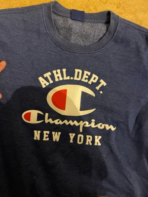 Blå sweatshirt från Champion - Säljer en blå sweatshirt från Champion med tryck i vitt och rött på bröstet där det står 'ATHL. DEPT. Champion NEW YORK'. Tröjan har rund halsringning och långa ärmar. Perfekt för en sportig och avslappnad stil.