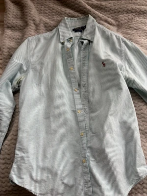 Randig skjorta från Polo Ralph Lauren - Snygg ljusblå och vit randig skjorta från Polo Ralph Lauren i slim fit. Klassisk button-down krage och broderad logga på bröstet. Tillverkad i mjuk bomull, perfekt för en clean och fräsch look.