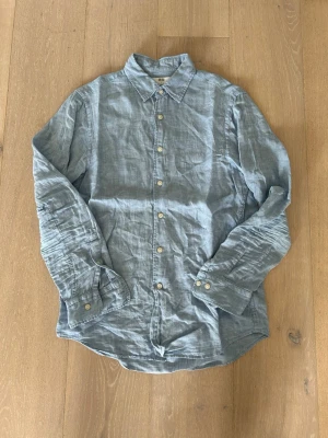 Linneskjorta från Uniqlo - Säljer en stilren ljusblå skjorta från Uniqlo i svalt linne. Skjortan har klassisk krage, långa ärmar och vita knappar framtill. Perfekt för dig som gillar en avslappnad men snygg look.