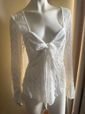 Vit spetsblus med knyt framtill strl XL - Snygg vit blus i spets med lång ärm och knyt framtill. Blusen har en djup v-ringning och är lätt transparentvid ärmar mage rygg ej vid byst. Den har ett romantiskt uttryck. Perfekt för dig som gillar feminina detaljer och trendig stil.