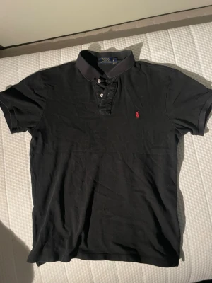 Svart pikétröja från Polo Ralph Lauren - Klassisk svart pikétröja från Polo Ralph Lauren med röd broderad logga på bröstet. Den är använd men inga defekter på. Väldigt bra till dig som vill ha en enkel och snygg pike med bra passform till sommaren! Strl M passar S