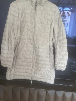 Ljusbeige lång dunjacka Parajumpers - Snygg ljusbeige lång dunjacka från Parajumpers med hög krage och dragkedja framtill. Jackan har ett emblem på ärmen och quiltad design som ger en modern look. Perfekt för kalla vinterdagar och riktigt varm tack vare dunfyllningen.