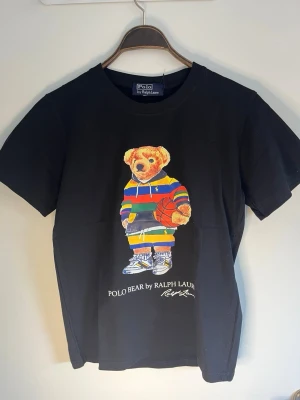 Svart Polo Bear t-shirt Ralph Lauren - Svart t-shirt från Polo Ralph Lauren med ikoniska Polo Bear-trycket framtill. Björnen har en färgglad randig hoodie och håller en basketboll. Klassisk rund hals och korta ärmar. Perfekt statement-plagg för dig som gillar streetstyle.