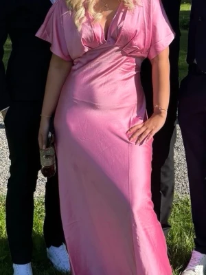 Rosa klänning -  rosa långklänning med djup v-ringning och korta ärmar. Klänningen har omlottdetalj vid bysten och ett mjukt, glansigt material som ger ett lyxigt intryck. Perfekt för speciella tillfällen och studentfirande. Använd en gång, inga defekter.