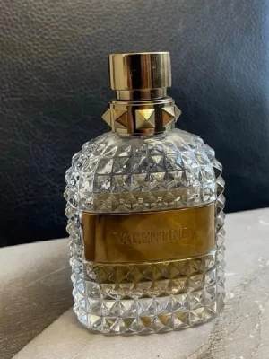 Valentino parfym med guldiga detaljer - Säljer en lyxig parfym från Valentino i en genomskinlig glasflaska med nitar och guldiga detaljer. Flaskan har ett elegant mönster med små pyramidformade nitar och en bred guldfärgad etikett. Lock och hals är också dekorerade med guldfärgade detaljer.