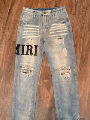 Amiri Jeans - Säljer ett par ljusblå jeans från Amiri med stora svarta bokstäver, slitningar och hål på framsidan samt distressed detaljer. Jeansen har raka ben, fem fickor och coola tvättade effekter. Perfekta för dig som gillar streetwear och unik stil.