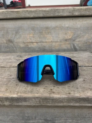 Blå sportiga solglasögon - Säljer ett par riktigt snygga sportiga solglasögon med spegelblått, kantigt glas och svart båge. De har en futuristisk vibe och passar perfekt för dig som vill sticka ut. Glasen är breda och ger bra täckning, och bågarna är i lätt syntetmaterial.