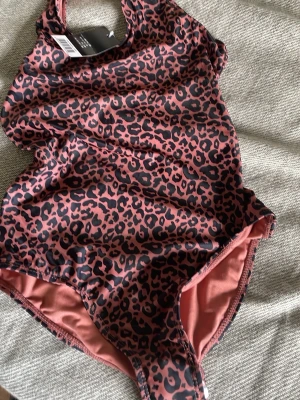 Leopardmönstrad baddräkt i rosa och svart - Snygg baddräkt med leopardmönster i rosa och svart. Klassisk modell med rund halsringning och breda axelband. Tillverkad i stretchigt material som sitter skönt på kroppen. Perfekt för dig som vill sticka ut på stranden eller vid poolen.