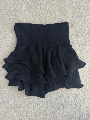 Svart volangkjol med hög midja - Supersnygg svart kjol med volanger och hög smockad midja. Kjolen har en kort modell och är perfekt för dig som gillar en feminin och trendig stil. Materialet känns lätt och flowy, vilket ger kjolen ett fint fall. Kan användes som en topp 