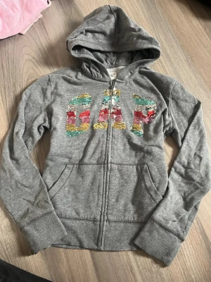 Grå hoodie med paljetter från GAP - Säljer en grå zip-hoodie från GAP med huva och stora fickor framtill. På bröstet står det GAP i stora bokstäver täckta av skimrande paljetter som skiftar färg mellan färgglatt och vitt beroende på hur man drar dem. Perfekt för dig som gillar lite extra bling i vardagen.