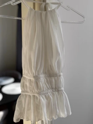Vit volang linnetopp med knyt - Vit linnetopp med volangkant och knytning i nacken och ryggen. Toppen är ärmlös och har en luftig, lätt rynkad design som ger en romantisk vibe. Perfekt för varma dagar och enkel att styla med jeans eller kjol. Endast testad.
