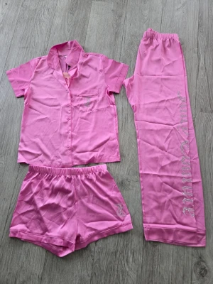 Rosa Juicy Couture pyjamas-set S  - Säljer ett rosa pyjamas-set från Juicy Couture i mjukt material. Setet består av en kortärmad skjorta med knappstängning, ett par shorts och ett par långa byxor. Både byxorna och shortsen har Juicy Couture-logga i strass. Perfekt för mysiga hemmakvällar!