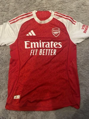 Arsenal matchtröja Adidas röd/vit M - Säljer en officiell elite player edition Arsenal matchtröja från Adidas i rött och vitt med klubbmärke och sponsortryck på bröstet. Tröjan har korta ärmar, rund hals och är tillverkad i ett lätt, ventilerande funktionsmaterial. Slim fit-modell, perfekt för fotboll eller träning. Kvitto finns