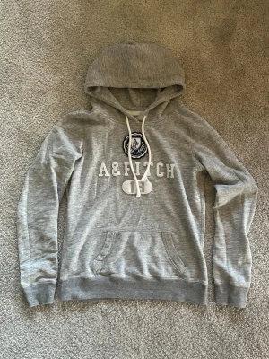 Grå hoodie från Abercrombie & Fitch - Snygg grå hoodie från Abercrombie & Fitch med vit broderad logga och text på bröstet. Klassisk känguruficka framtill, dragsko i huvan och ribbade muddar. Perfekt för en avslappnad och trendig look. Mjuk bomullsblandning för extra komfort.