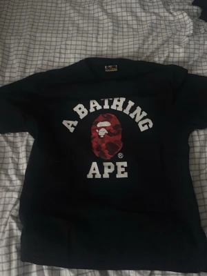 Svart A Bathing Ape t-shirt - Säljer en svart t-shirt från A Bathing Ape med det klassiska BAPE-logotrycket i vitt och en röd kamouflageapa på bröstet. T-shirten har rund hals och korta ärmar. Perfekt för dig som gillar streetwear och vill sticka ut. Tröjam känd inte som L utan skulle mer säga M