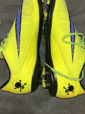 Nike hypervenom storlek 41 - Säljer ett par riktigt snygga fotbollsskor från Nike i gul och svart med cool dödskalle-detalj på hälen. Skorna har gröna snören, svart sula med gula dobbar och en blå/svart detalj på ovansidan. Perfekta för dig som vill sticka ut på planen!
