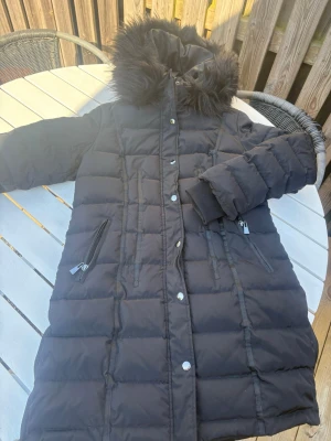 Svart lång dunjacka med fuskpäls - Snygg svart lång dunjacka med quiltad design och stor huva med fuskpäls. Jackan har dragkedja och tryckknappar framtill samt två snedställda fickor med dragkedja. Perfekt för kalla vinterdagar och riktigt mysig att bära.