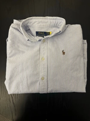 Randig skjorta från Polo Ralph Lauren - Klassisk skjorta från Polo Ralph Lauren i ljusblått och vitt med tunna ränder. Skjortan har lång ärm, knappar framtill och den ikoniska broderade loggan på bröstet. normal passform.