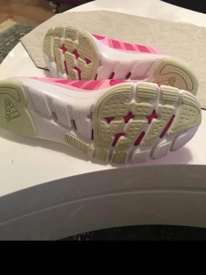 Rosa Adidas sneakers med meshdetaljer - Snygga rosa Adidas sneakers med vita och ljusgröna detaljer. Skorna har en låg profil, klassiska tre ränder på sidan och ovandel i mesh för extra andningsförmåga. Yttersulan är vit med ljusgröna och rosa inslag. Perfekta för en sportig och trendig look.