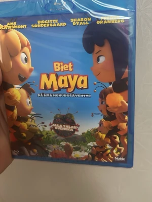 Biet Maya Blu-ray film - Säljer Blu-ray-filmen 'Biet Maya: På nya honungsäventyr'. Omslaget är blått med färgglada illustrationer av Maya och hennes vänner. Alla talar svenska och filmen är animerad. Perfekt för dig som gillar tecknade äventyr och familjefilmer.