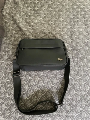 Svart axelväska från Lacoste - Snygg svart axelväska från Lacoste med justerbar axelrem och silverfärgad krokodillogo på framsidan. Väskan har en rektangulär form, ytterfack med dragkedja och är tillverkad i ett tåligt konstlädermaterial. Perfekt för att bära det viktigaste med stil.