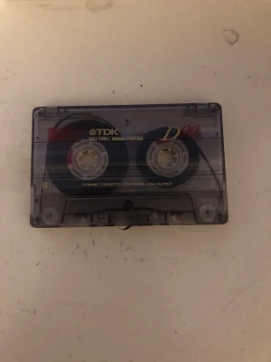 TDK D90 kassettband - Klassiskt TDK D90 kassettband i transparent plast med svarta och röda detaljer. Bandet har tydliga A- och B-sidor och rymmer 90 minuter ljud. Perfekt för dig som gillar retroprylar eller vill spela in egen musik.