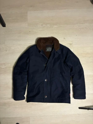 Marinblå Levi's jacka med teddyfoder - Snygg marinblå jacka från Levi's med brunt teddyfoder och krage. Jackan har två sidofickor, knäppning framtill och en klassisk, rak passform. Perfekt för kyligare dagar när du vill ha både stil och värme.