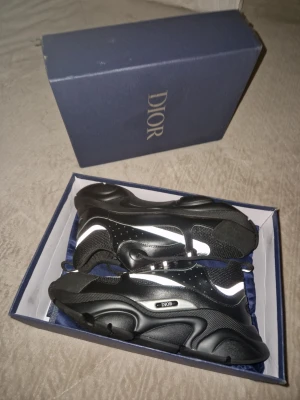 Svarta Dior sneakers med vita detaljer - Snygga svarta Dior sneakers med vita reflekterande detaljer och chunky sula. Skorna har en modern design med mesh och läderpaneler, samt logotyp på sidan. Perfekta för dig som vill ha en exklusiv och trendig look.