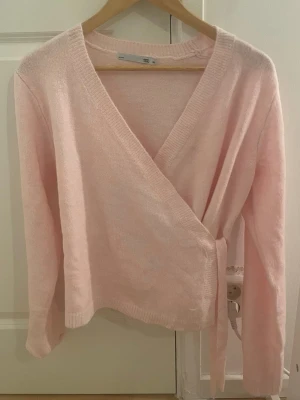 Ljusrosa omlottkofta med knytband - Supersöt ljusrosa stickad omlottkofta med långa ärmar och knytband i midjan. Koftan har en mjuk och tunn stickning som ger en avslappnad men ändå feminin look. Perfekt att slänga över en topp för extra lager.