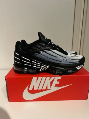 Nike Air Max Plus 3 svart/vit - Nike Air Max Plus 3 sneakers i svart och vitt med gradient-effekt. Skon har en futuristisk design med synliga Air Max-bubblor i sulan, svarta detaljer och vita linjer på sidan. Ovandelen är i mesh och syntetmaterial, och tungan har TN Air Max-logga.