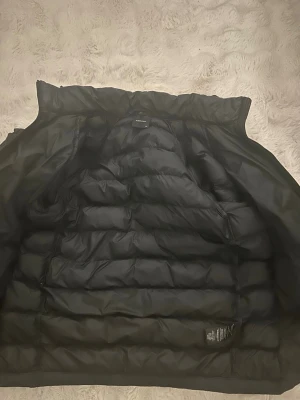 Svart dunjacka från Peak Performance - Säljer en svart dunjacka från Peak Performance med huva och dragkedja framtill. Jackan har två sidofickor med dragkedja och diskret logga på bröstet. Puffig design med breda kanaler och varm vaddering, perfekt för kalla vinterdagar.
