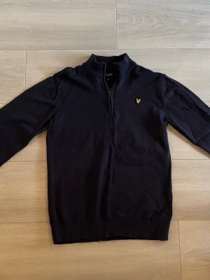 Mörkblå halvzip från Lyle & Scott - Mörkblå tröja med halv dragkedja från Lyle & Scott. Klassisk modell med lång ärm och gul logga broderad på bröstet. Tillverkad i mjukt material som känns skönt mot huden. Perfekt till jeans eller chinos för en clean look.