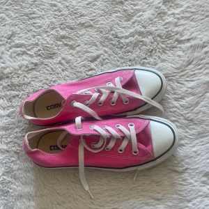 Rosa Converse sneakers låga - Säljer ett par klassiska låga Converse sneakers i stark rosa färg med vita snören och tåhätta. Skorna har vit sula med svart rand och är tillverkade i canvasmaterial. Perfekta för dig som vill sticka ut med färg på fötterna.