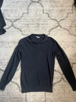 Mörkblå stickad tröja John Henric - Mörkblå stickad tröja från John Henric med klassisk krage och lång ärm. Tröjan har en stilren design utan mönster och är tillverkad i ett mjukt, tunt material som passar perfekt till lager på lager. Ribbstickade muddar vid ärmslut och nederkant.